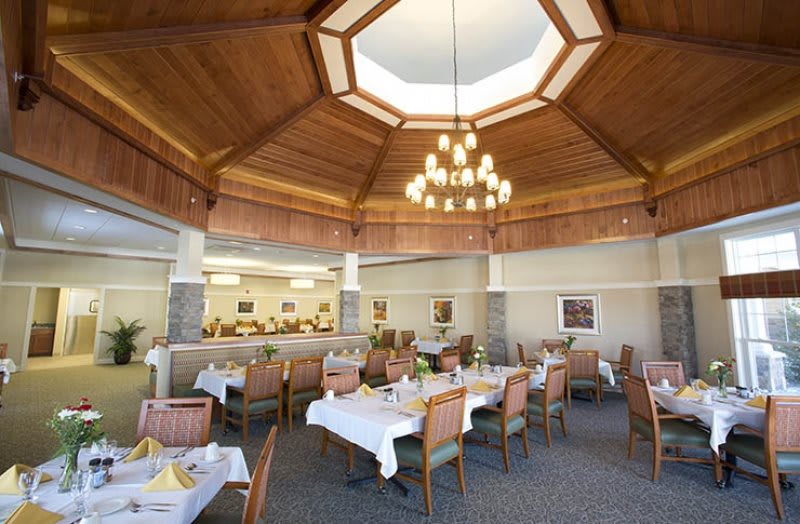 A well-lit restaurant interior with a stylish chandelier, featuring neatly arranged tables and chairs under a wooden ceiling.