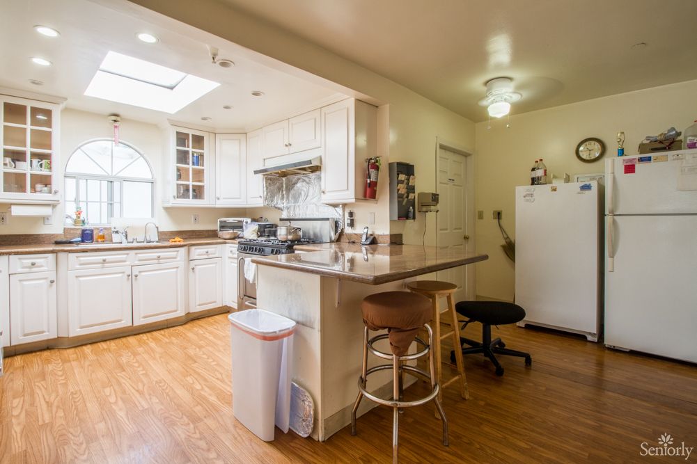 A modern kitchen interior with white cabinets and wood flooring, featuring a large countertop with bar stools.