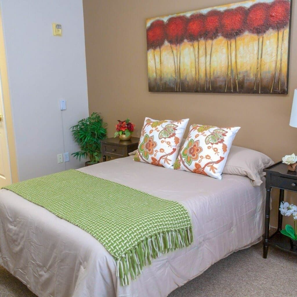 A cozy bedroom with a neatly made bed, adorned with floral pillows and a green throw, complemented by wall art and a bedside lamp.