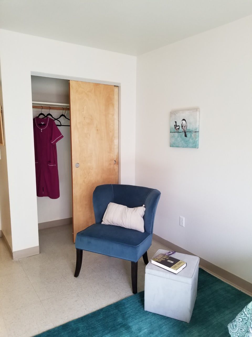 A cozy room with a blue chair near an open closet, featuring a dress and a small table with a book.