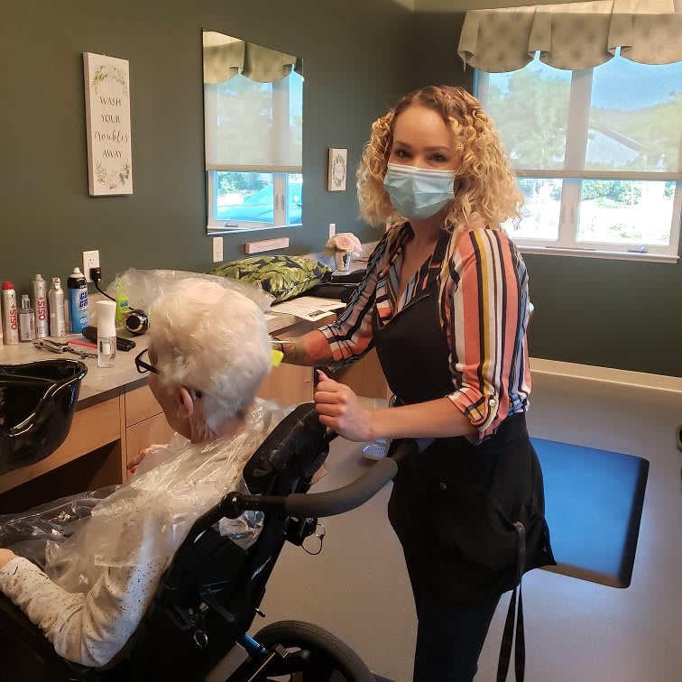 A hairdresser wearing a mask styles the hair of an elderly person in a salon.