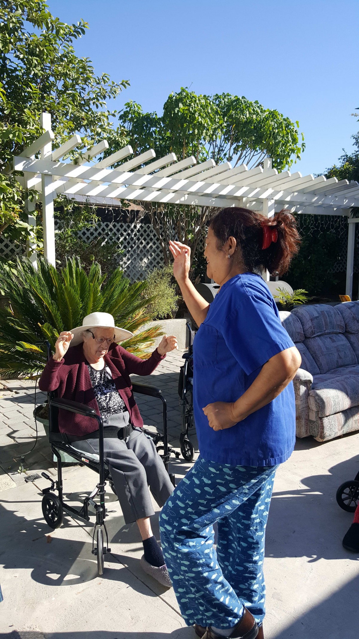 A caregiver dances outdoors near a person in a wheelchair on a sunny day.