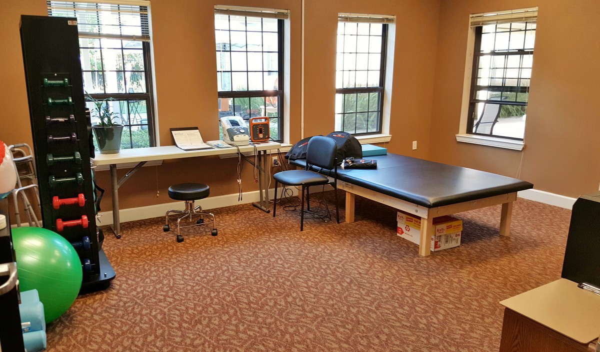 A therapy treatment room with a table, chairs, and medical equipment against the backdrop of large windows.