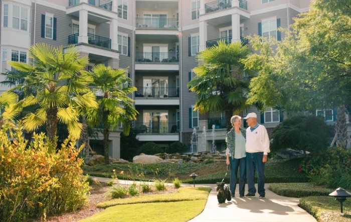 An elderly couple with a dog stands in front of a modern apartment building surrounded by palm trees and a garden.