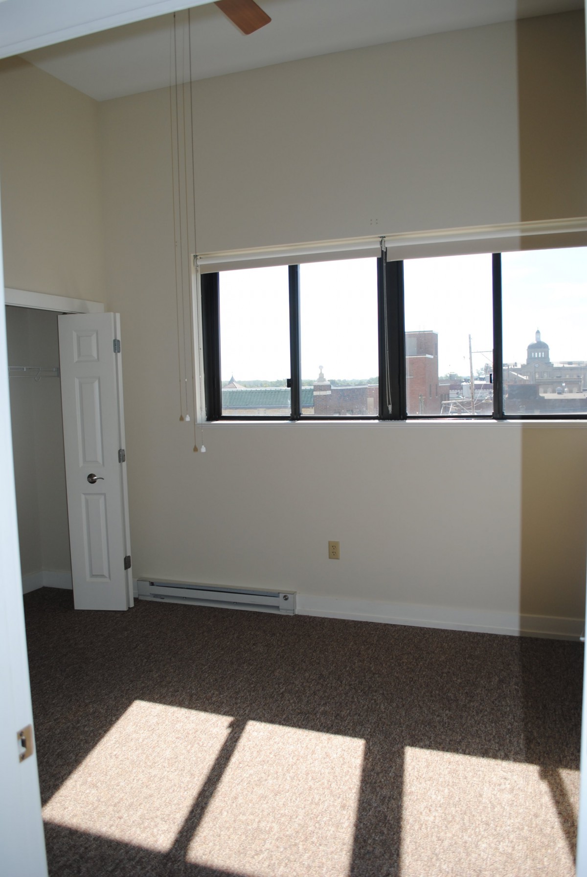 An empty room with carpet flooring and a large window letting in sunlight.