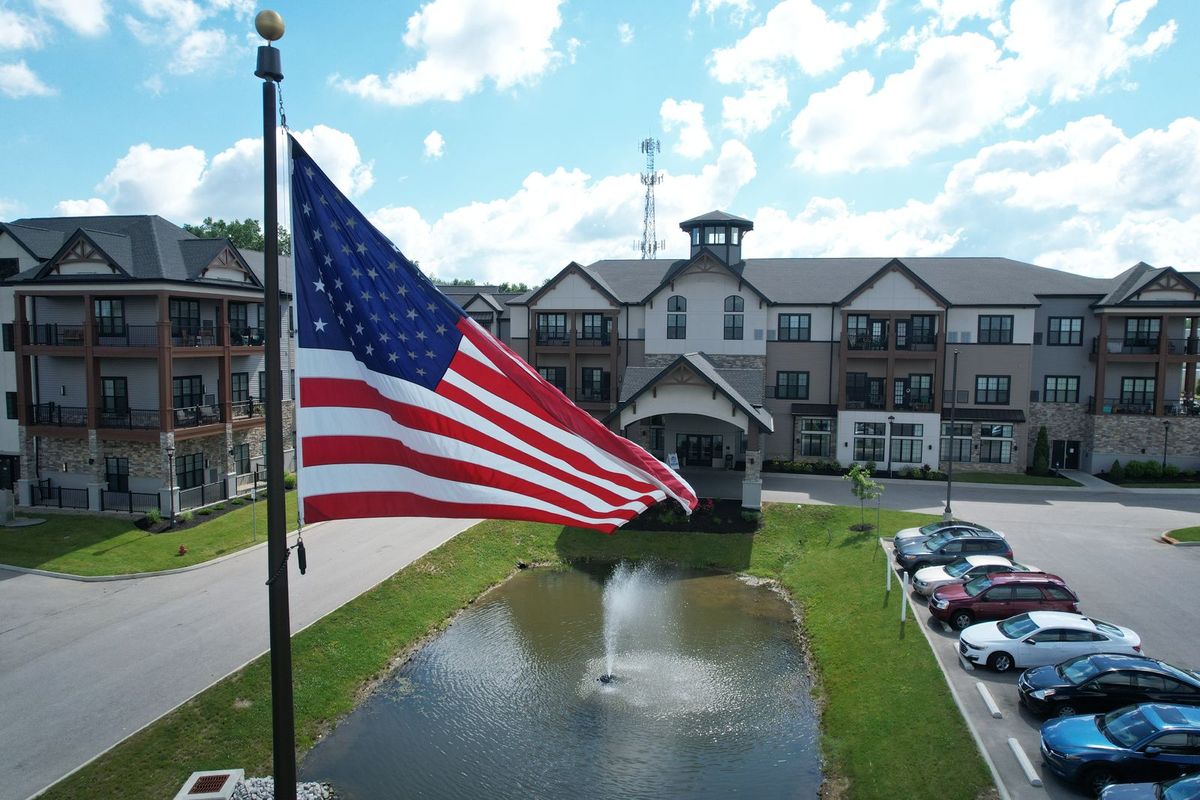 An American flag waves in front of a large building with a pond and fountain, set against a cloudy sky.