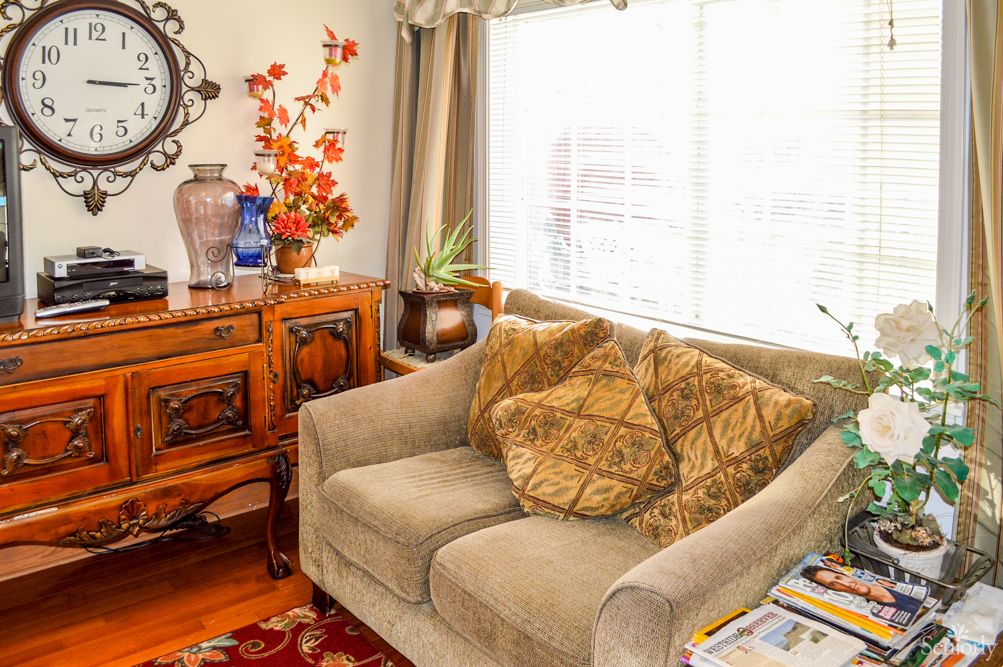 A cozy living room with a beige couch, decorative pillows, and a sunlit window. A wooden cabinet with vases and plants adds charm.