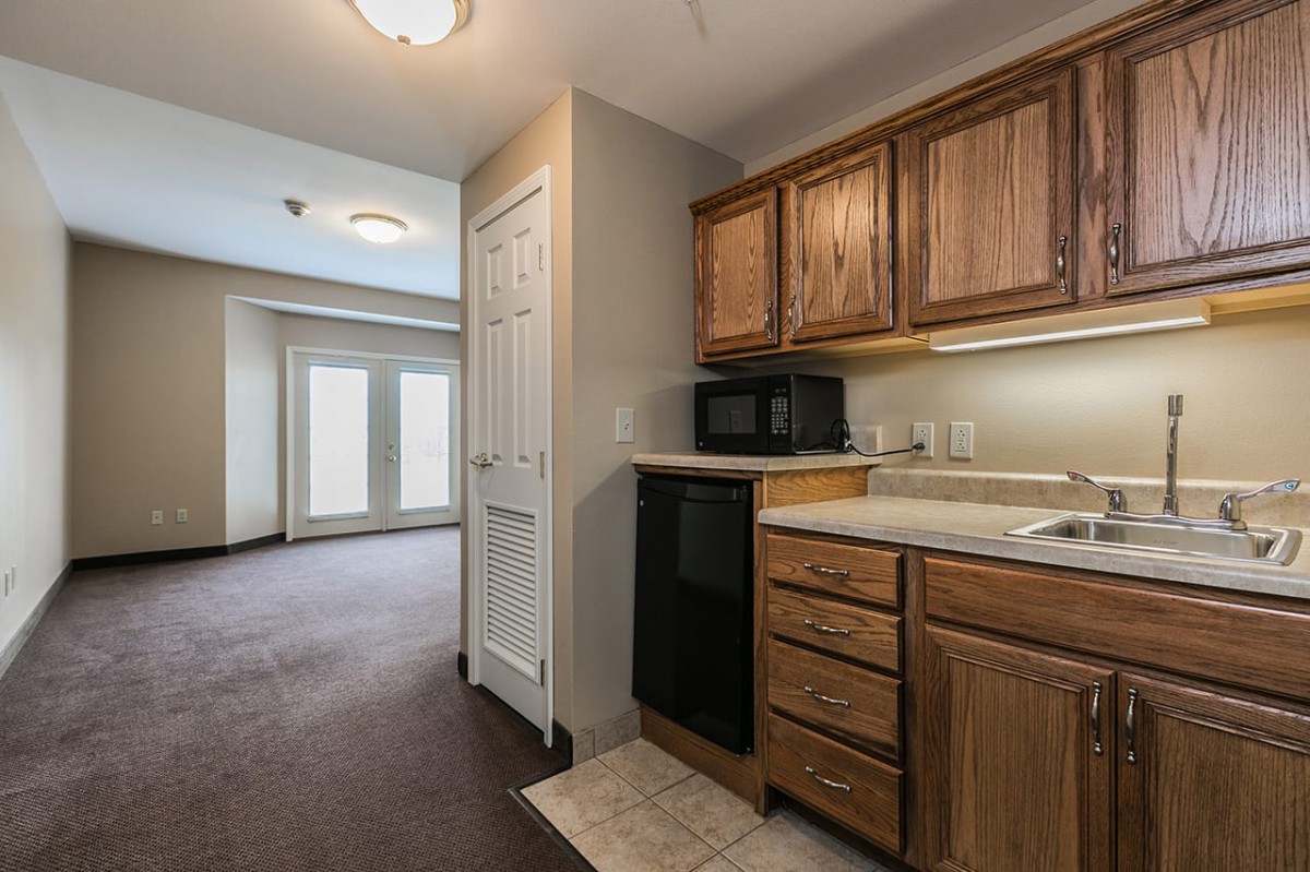 A view of a small kitchenette with wooden cabinets and a microwave, leading to a living area with large doors.