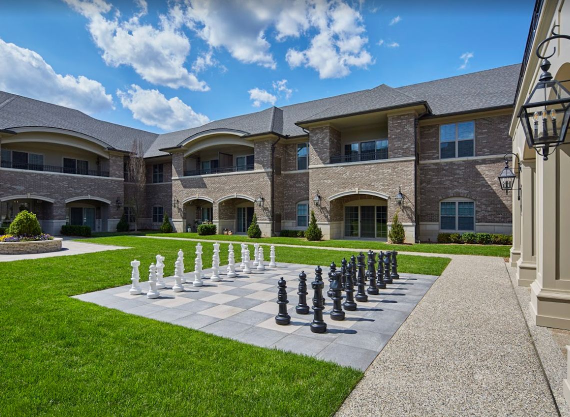 A large outdoor chessboard with oversized chess pieces sits in front of a brick building on a sunny day.