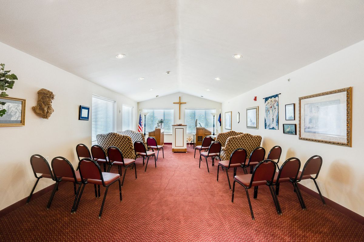 A small chapel interior with rows of chairs facing a pulpit and cross, featuring a red carpet and large windows.