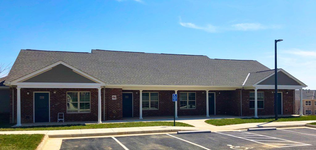 A single-story brick residential building with a sloped roof and an adjacent parking lot under a clear blue sky.