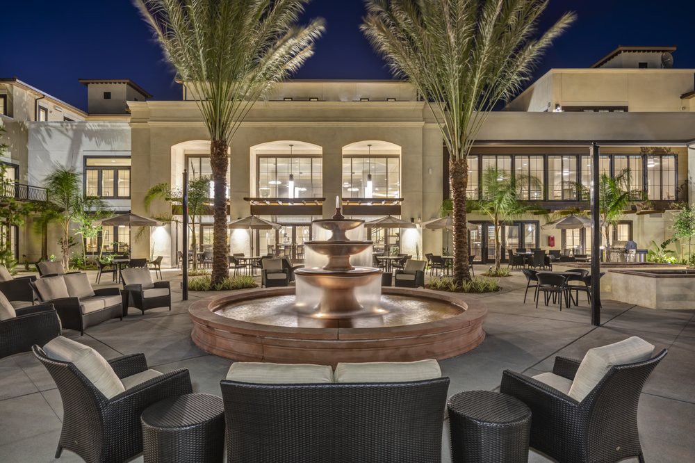 A luxurious outdoor setting with a central fountain surrounded by seating and palm trees, illuminated at night.