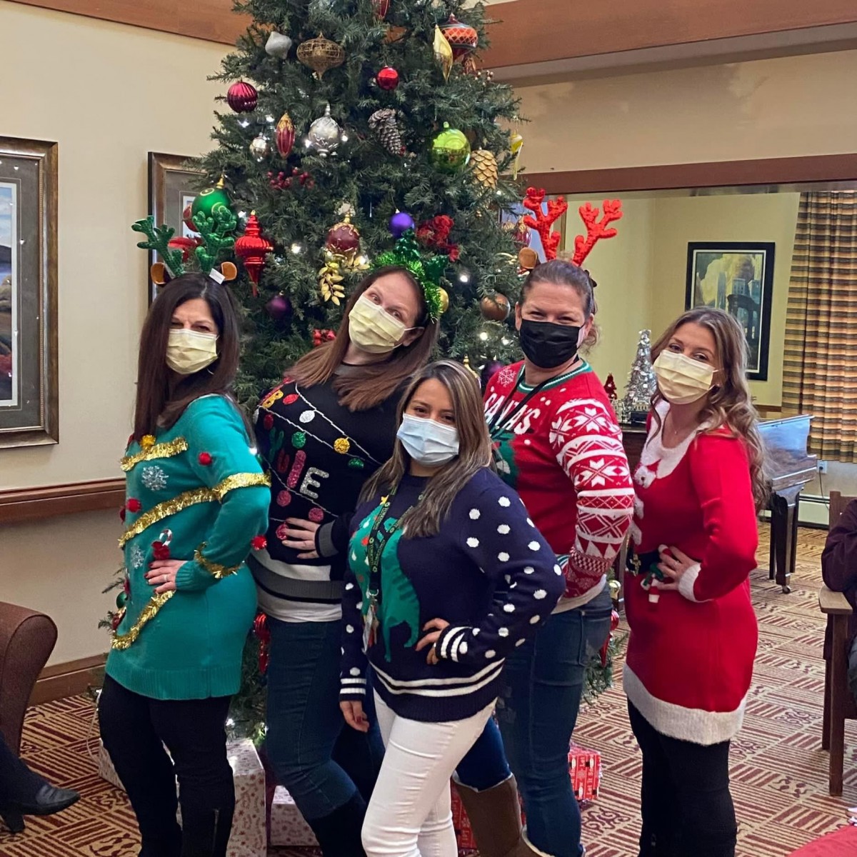 A group of people wearing festive holiday sweaters and masks pose in front of a decorated Christmas tree.