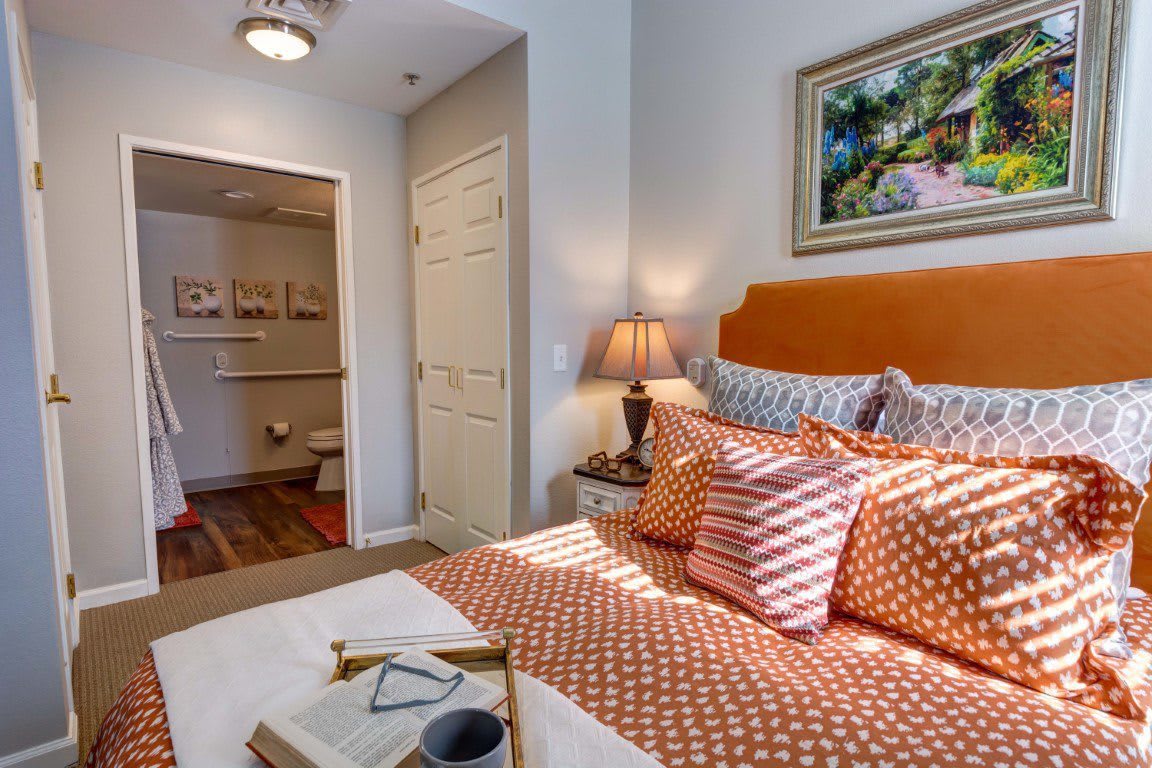 A cozy bedroom featuring a neatly made bed with patterned pillows, a side lamp, and artwork on the wall.