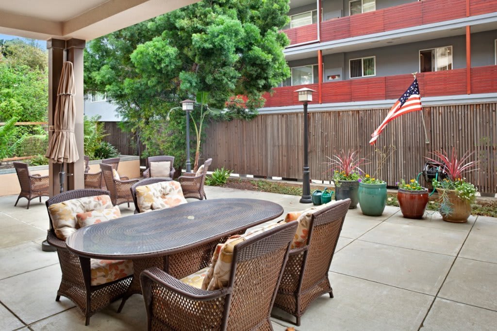 A cozy patio with wicker furniture and a dining table, surrounded by greenery and an American flag in the background.