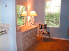 A cozy bedroom corner featuring a dresser with a mirror, adorned by lamps and decorative items, next to a window and a small chair.