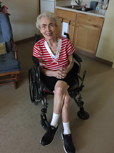 An elderly woman sitting in a wheelchair, wearing a striped shirt and smiling warmly.