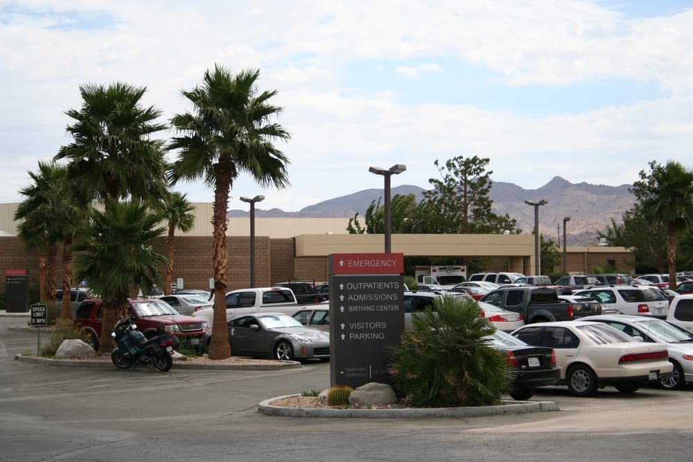 A hospital with a busy parking lot, surrounded by palm trees and mountains in the background.