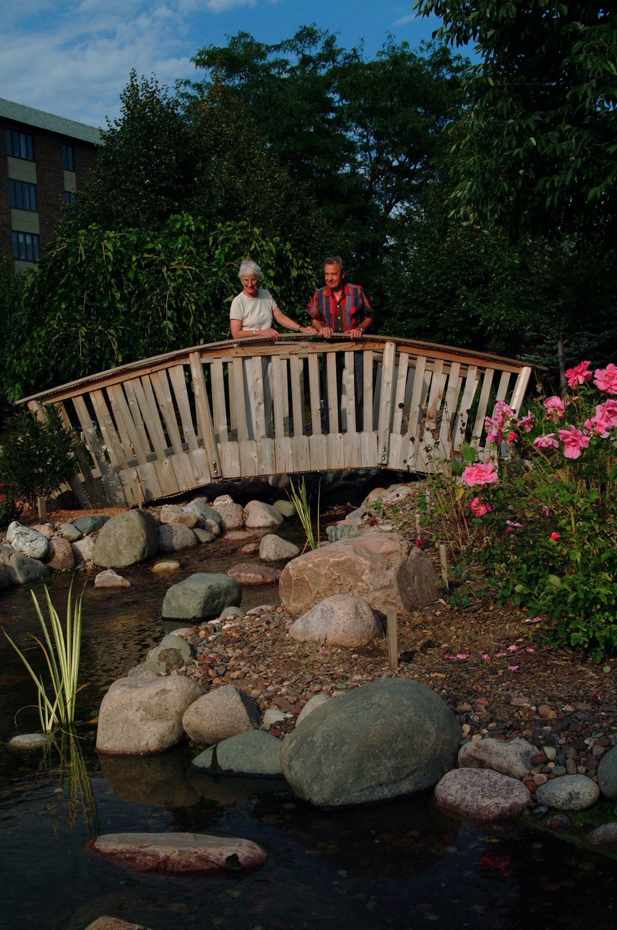 A couple standing on a wooden bridge over a garden pond surrounded by rocks and flowers.