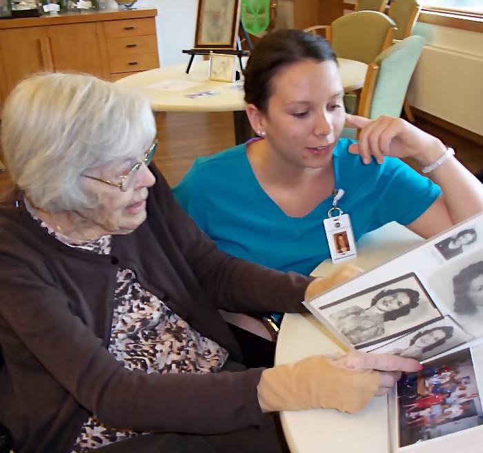 An elderly woman and a caregiver sitting at a table, looking through a photo album together.