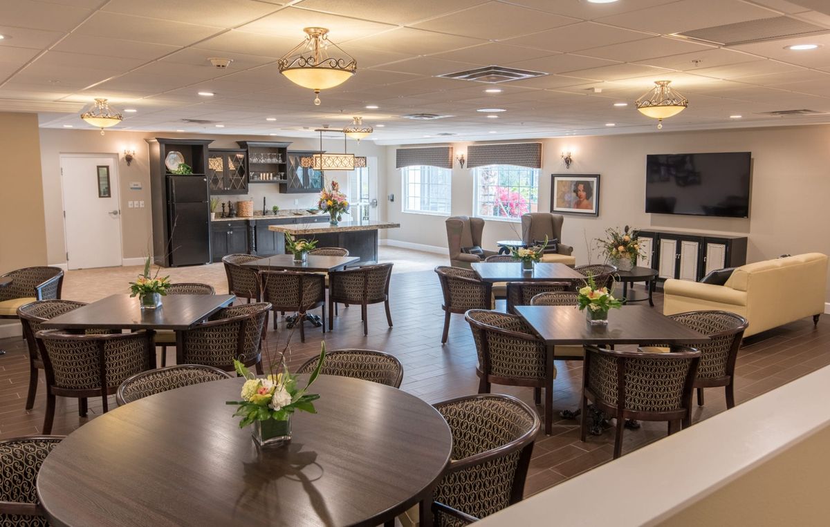 A well-lit dining area with multiple tables and chairs, featuring a kitchen in the background and decorative flowers on the tables.