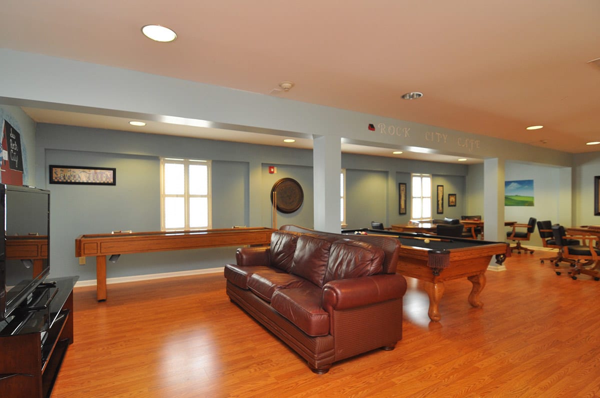 A spacious game room featuring a pool table, shuffleboard, and a comfortable leather sofa on a hardwood floor.