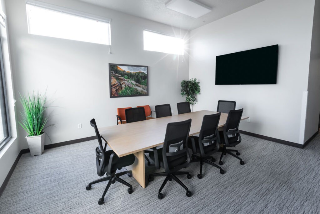 A modern conference room with a long table, several office chairs, a television on the wall, and decorative elements such as a potted plant and framed artwork.
