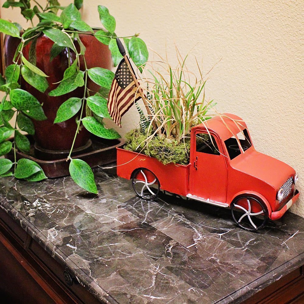 A charming decorative display featuring a red miniature truck with plants and an American flag on a marble surface.