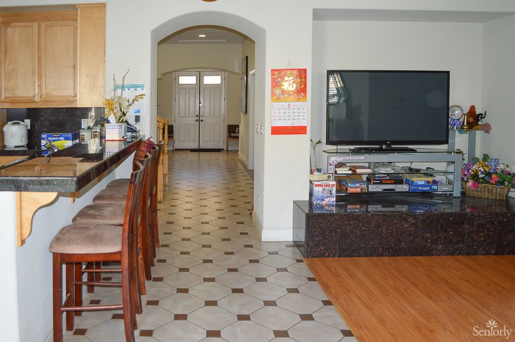A modern kitchen with bar stools leads into a hallway, adjacent to a living area featuring a large television.