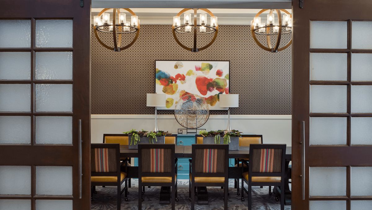 A stylish dining room featuring a large table with chairs, under elegant chandeliers and colorful artwork on the wall.