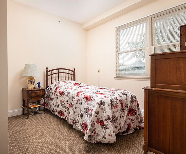 A cozy bedroom with a single bed covered in a floral bedspread, accompanied by a nightstand and lamp next to the window.