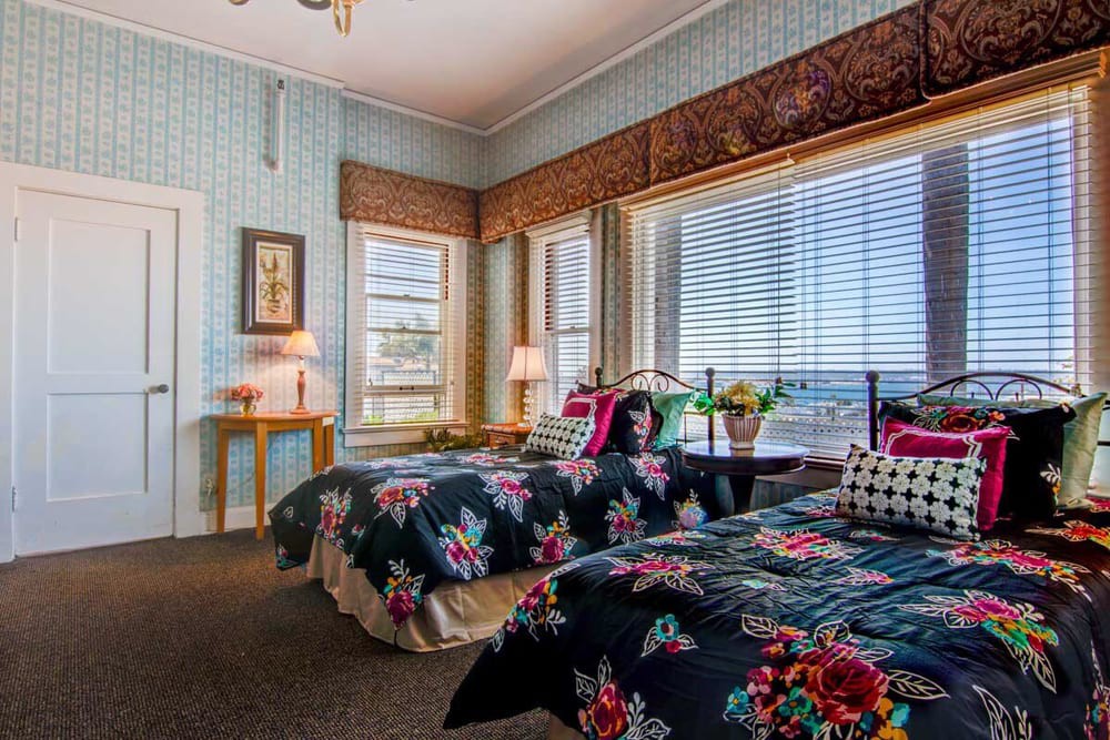 A cozy bedroom featuring twin beds with floral bedding, adorned with a large window allowing ample natural light.