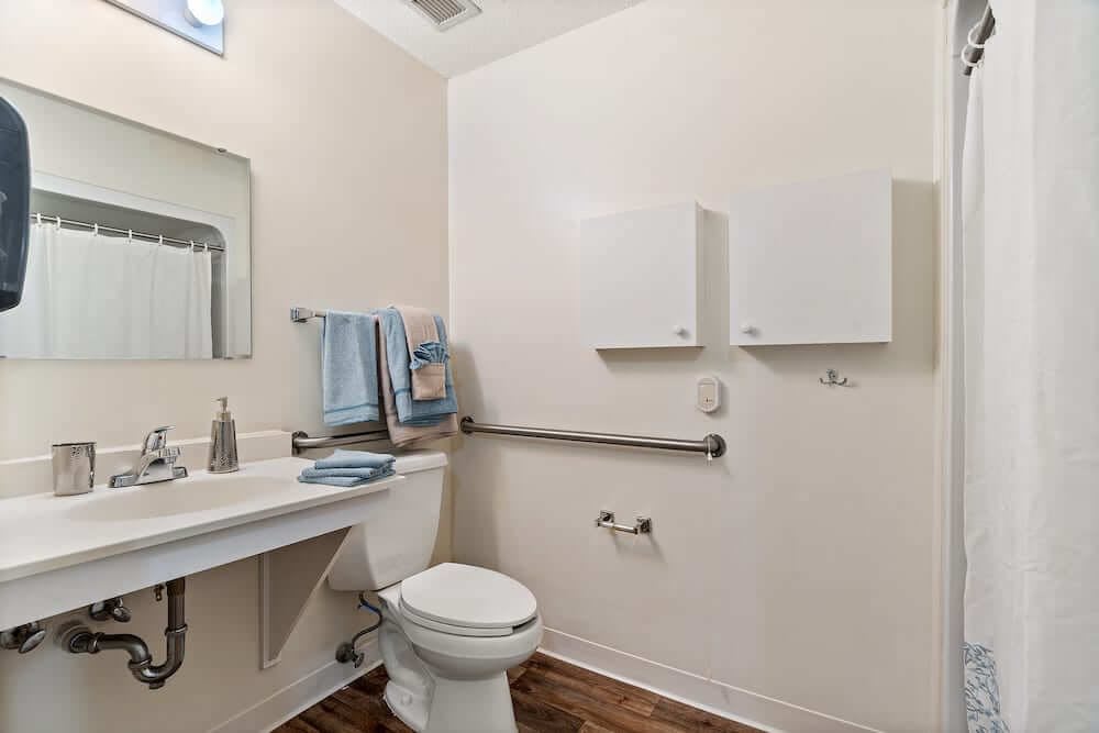 An accessible bathroom featuring a toilet, sink with mirror, towels, and handrails.