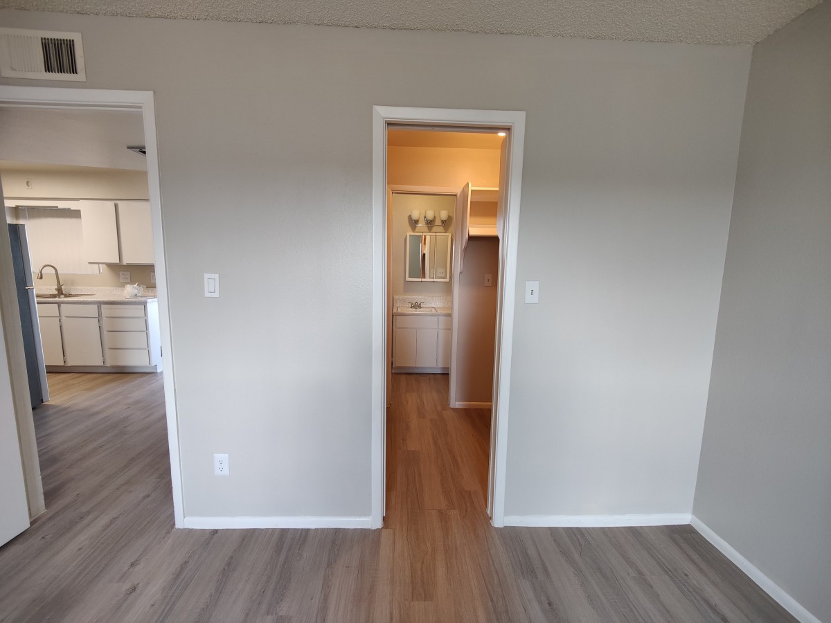 An empty room with a doorway leading to a hallway, featuring wooden flooring and white walls.