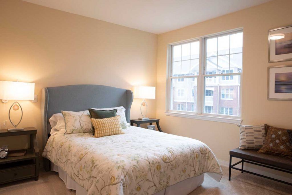 A cozy bedroom with a neatly made bed, bedside lamp, and large window allowing natural light.