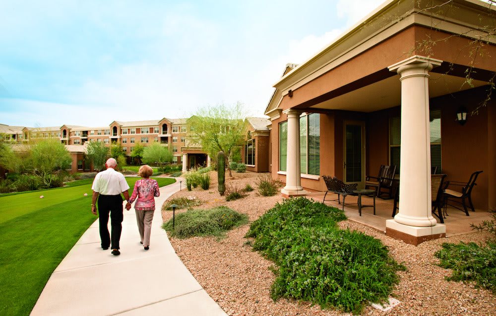An elderly couple walks along a garden path outside a retirement home building.