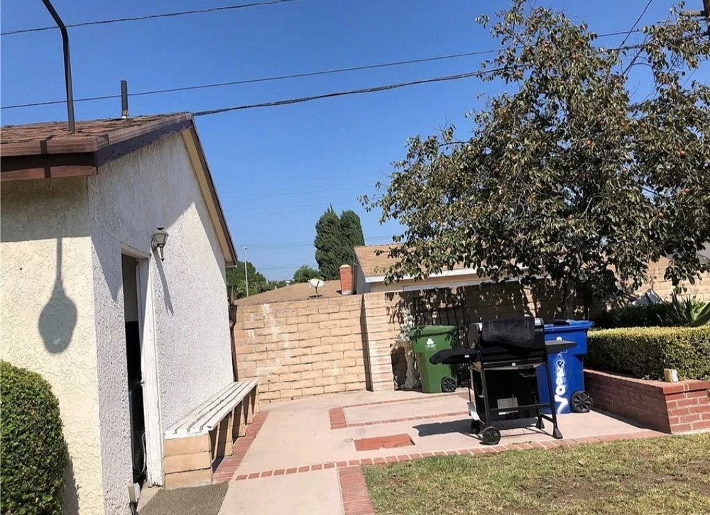 A sunny backyard patio area with a grill and recycling bins, surrounded by a tree and a house.