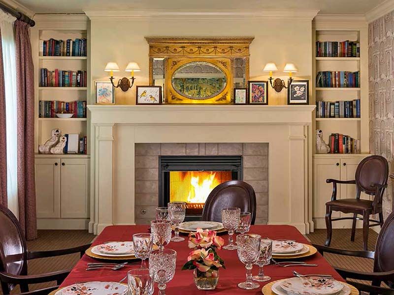 A cozy dining room with a beautifully set table in front of a fireplace, surrounded by bookshelves.