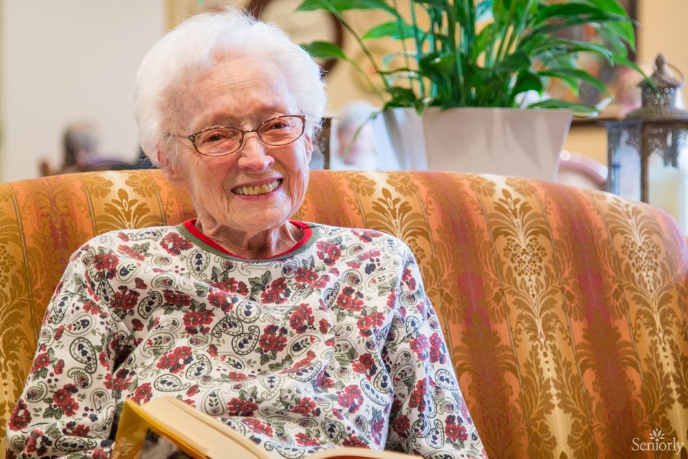 An elderly woman smiling while reading a book indoors.