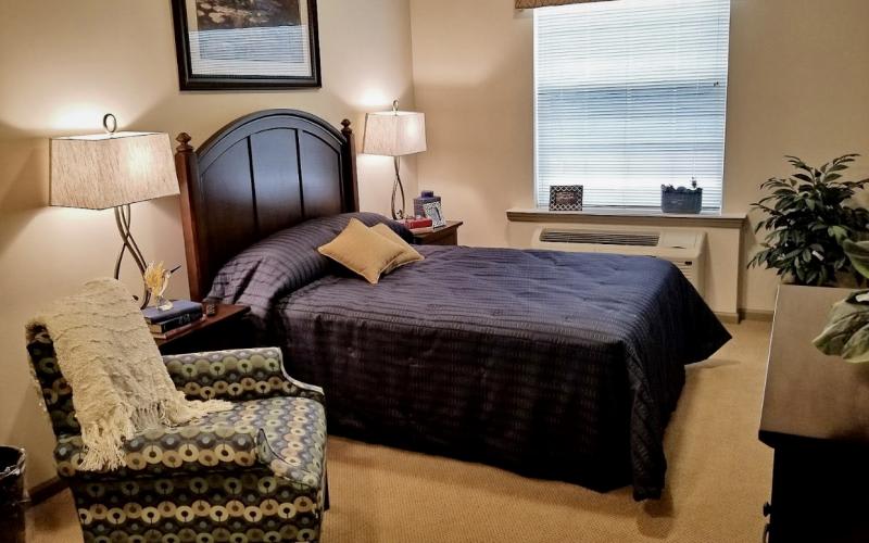 A cozy bedroom featuring a neatly made bed with a dark comforter, adjacent to a patterned armchair and a bedside lamp.