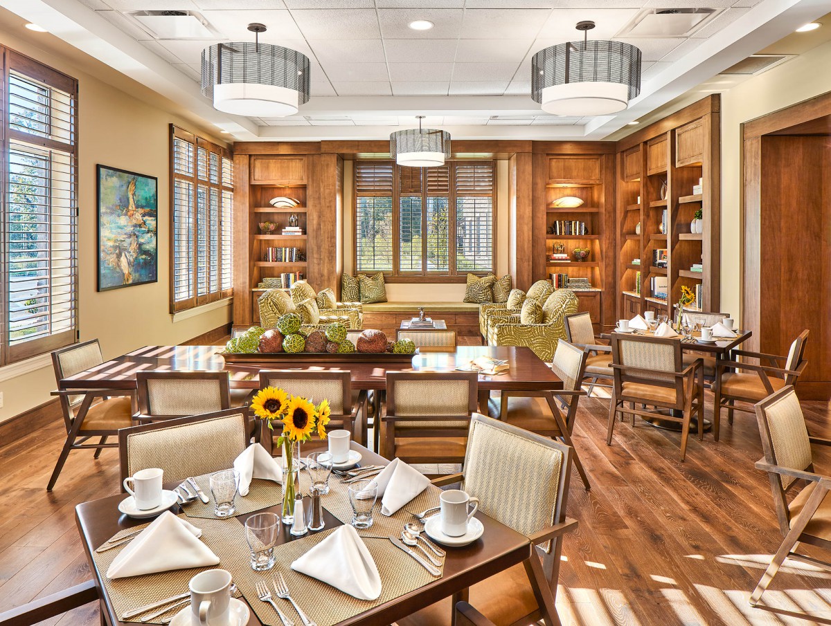 A cozy dining area with elegant interior design, featuring wooden floors, tables set for dining, comfortable seating, and a library section.