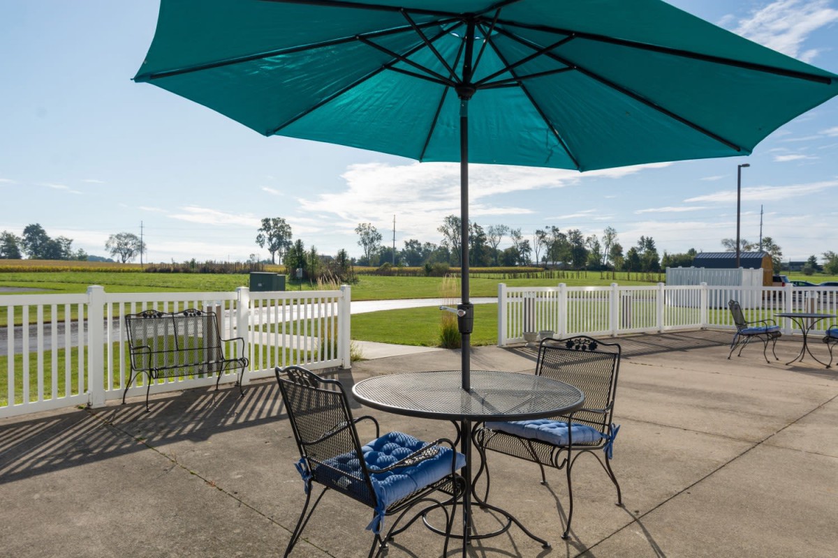 A sunny outdoor patio with a table, chairs, and a teal umbrella overlooking a landscaped area.