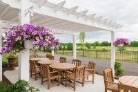 A charming outdoor patio with wooden tables and chairs under a white pergola, adorned with vibrant hanging flowers, overlooking a lush garden.