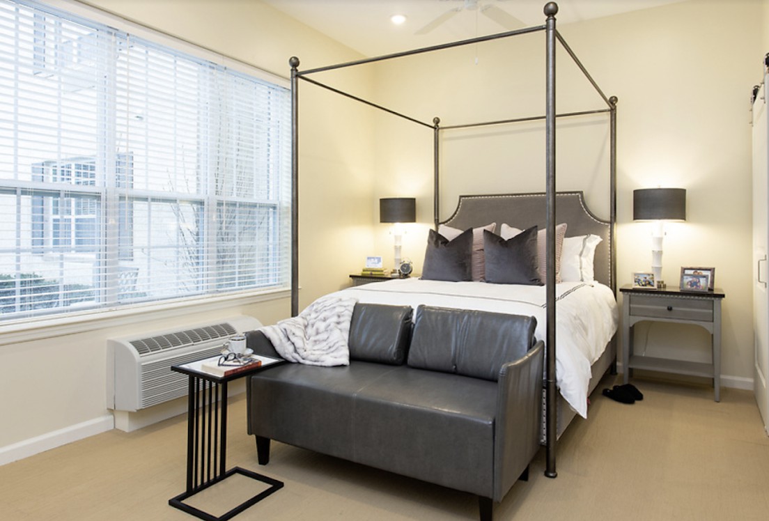 A modern bedroom featuring a canopy bed with elegant bedding, complemented by a gray sofa at the foot and stylish table lamps on each side.