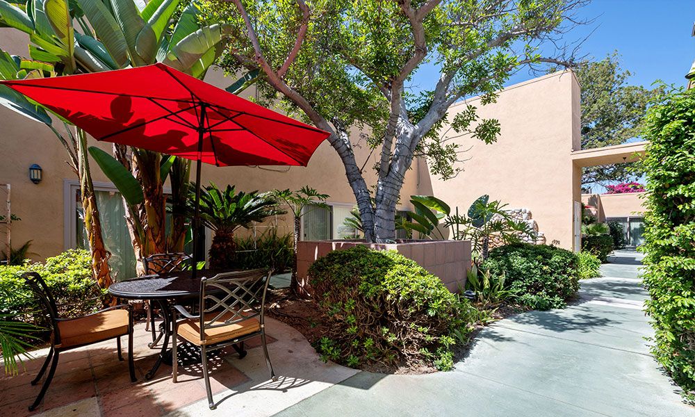 A cozy outdoor patio with a red umbrella, surrounded by lush greenery and a sunlit ambiance.