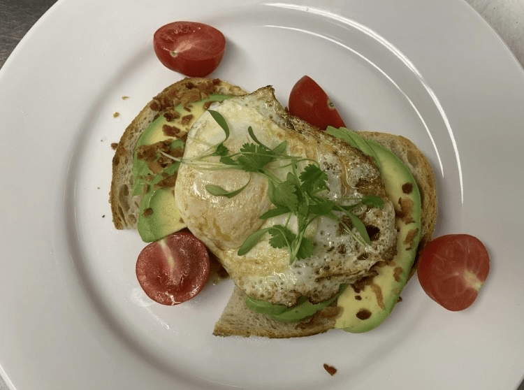 Avocado toast topped with a fried egg, garnished with herbs and served with cherry tomatoes.