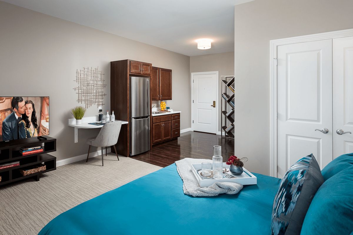 A modern apartment room with a kitchenette and a neatly arranged bed.