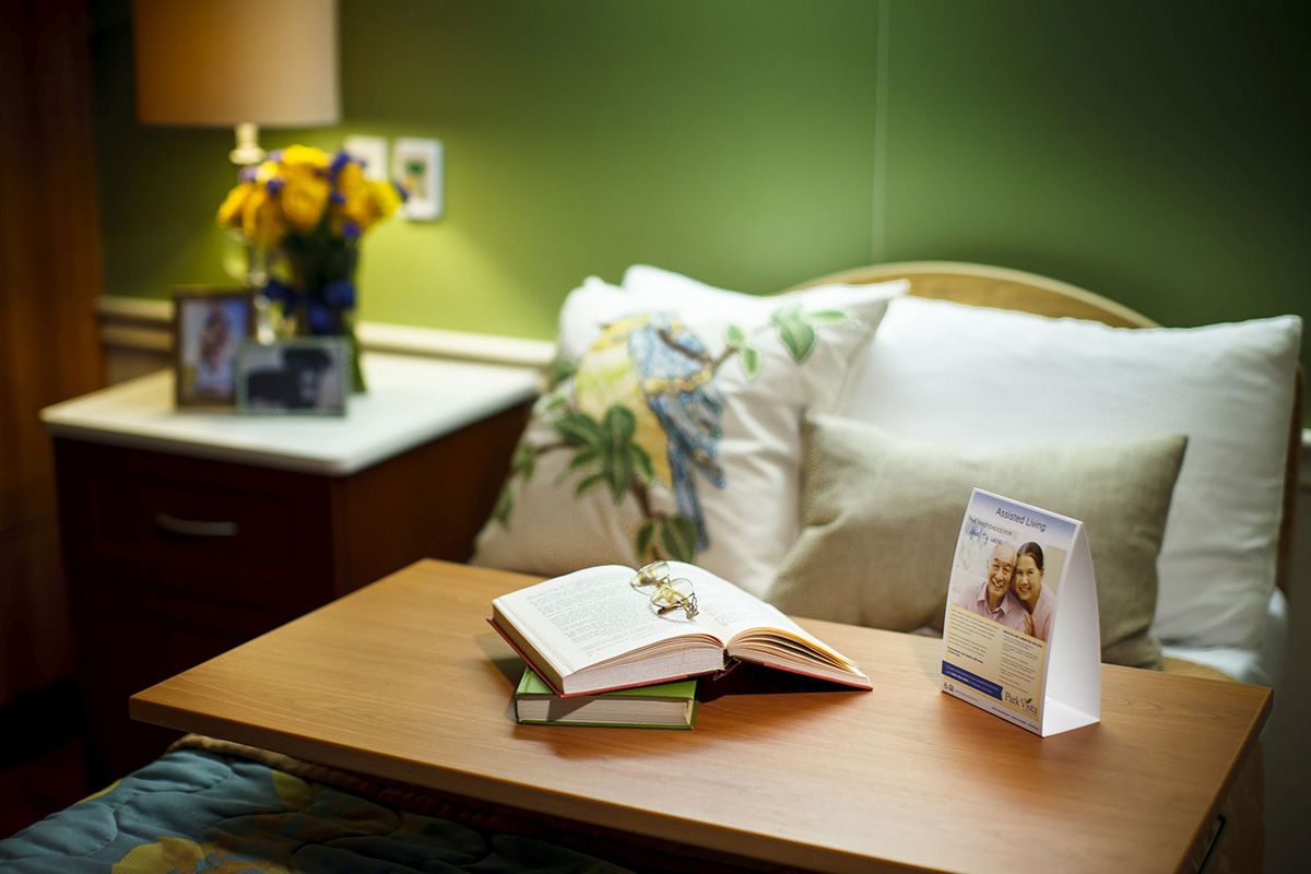 A cozy bedroom setup with an open book on a wooden table, surrounded by plush pillows and a bright bouquet of flowers under warm lighting.
