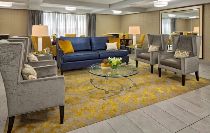 A modern living room with a blue sofa, gray chairs, a glass coffee table, and a patterned carpet.