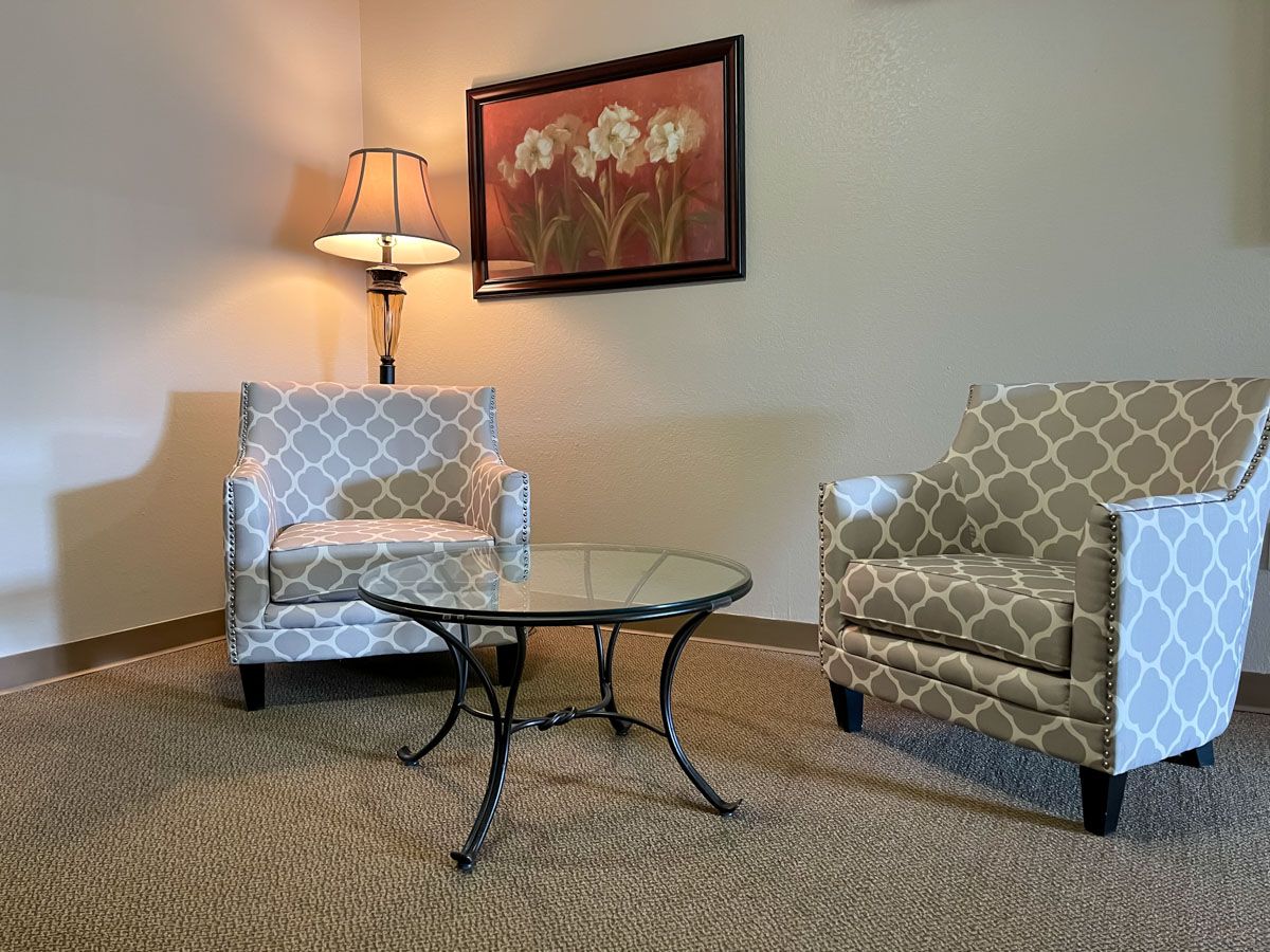 A cozy living room corner with patterned chairs and a glass-top table under warm lighting.